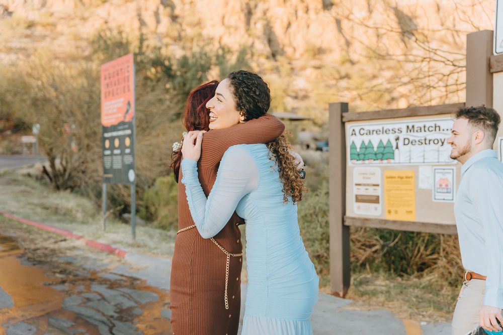 People share warm hugs and celebrations near a mountain trail marker.