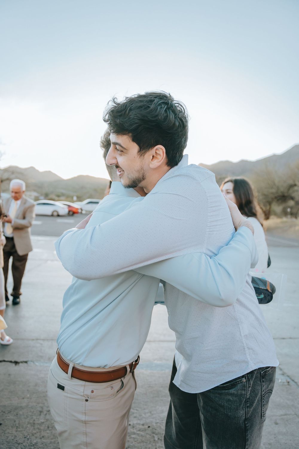 Two people in formal attire embrace and dance together at an outdoor desert location during sunset.
