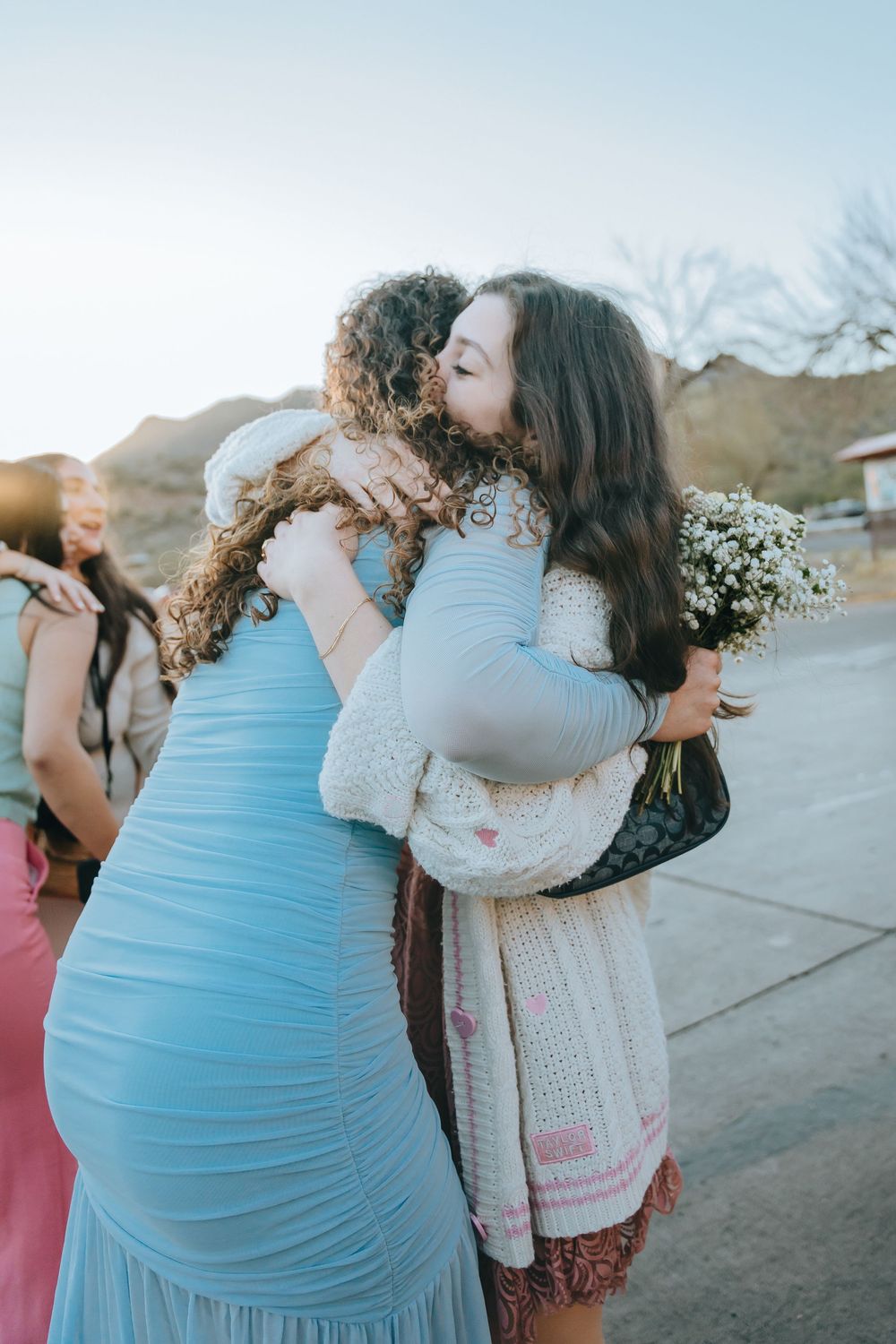 People share emotional embraces at an outdoor gathering with mountains in the background.