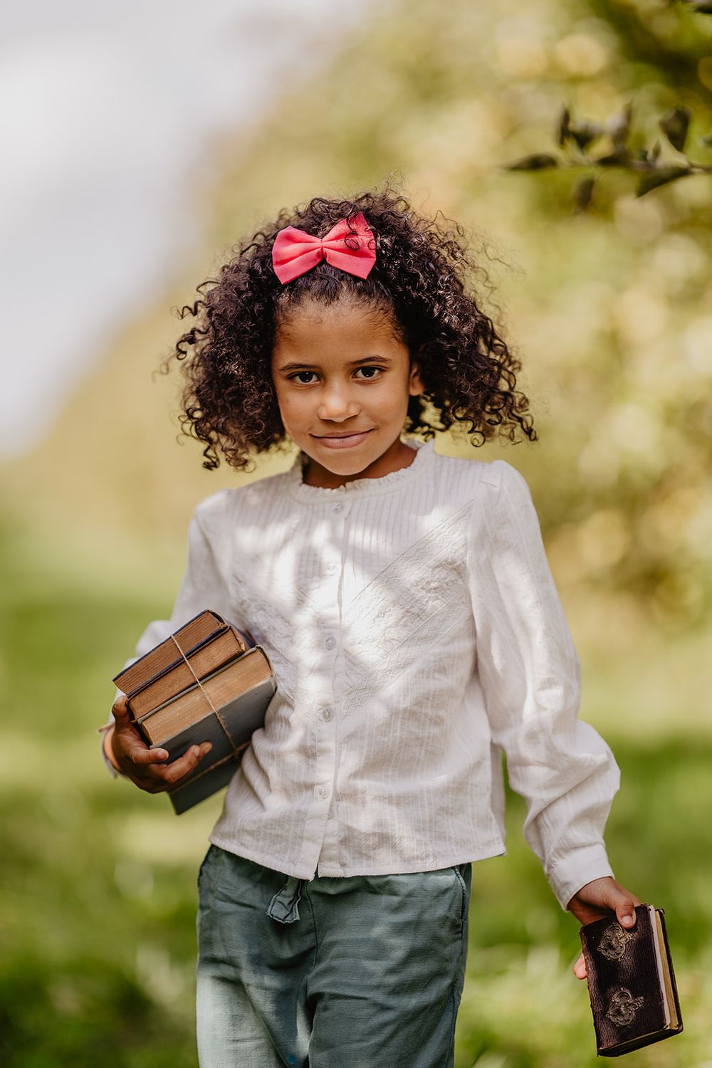 A young girl with curly hair and pink bow holds books while standing outdoors in a sunlit green garden.