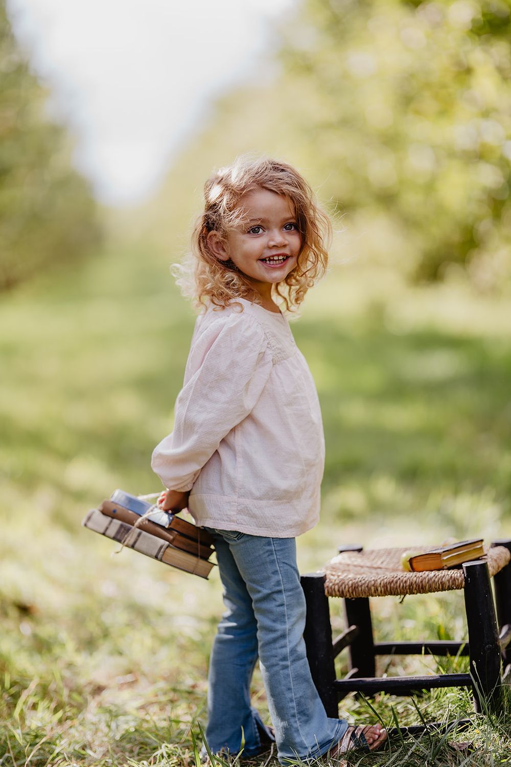 A cheerful curly-haired girl in jeans holds books while standing outdoors in a bright green garden.
