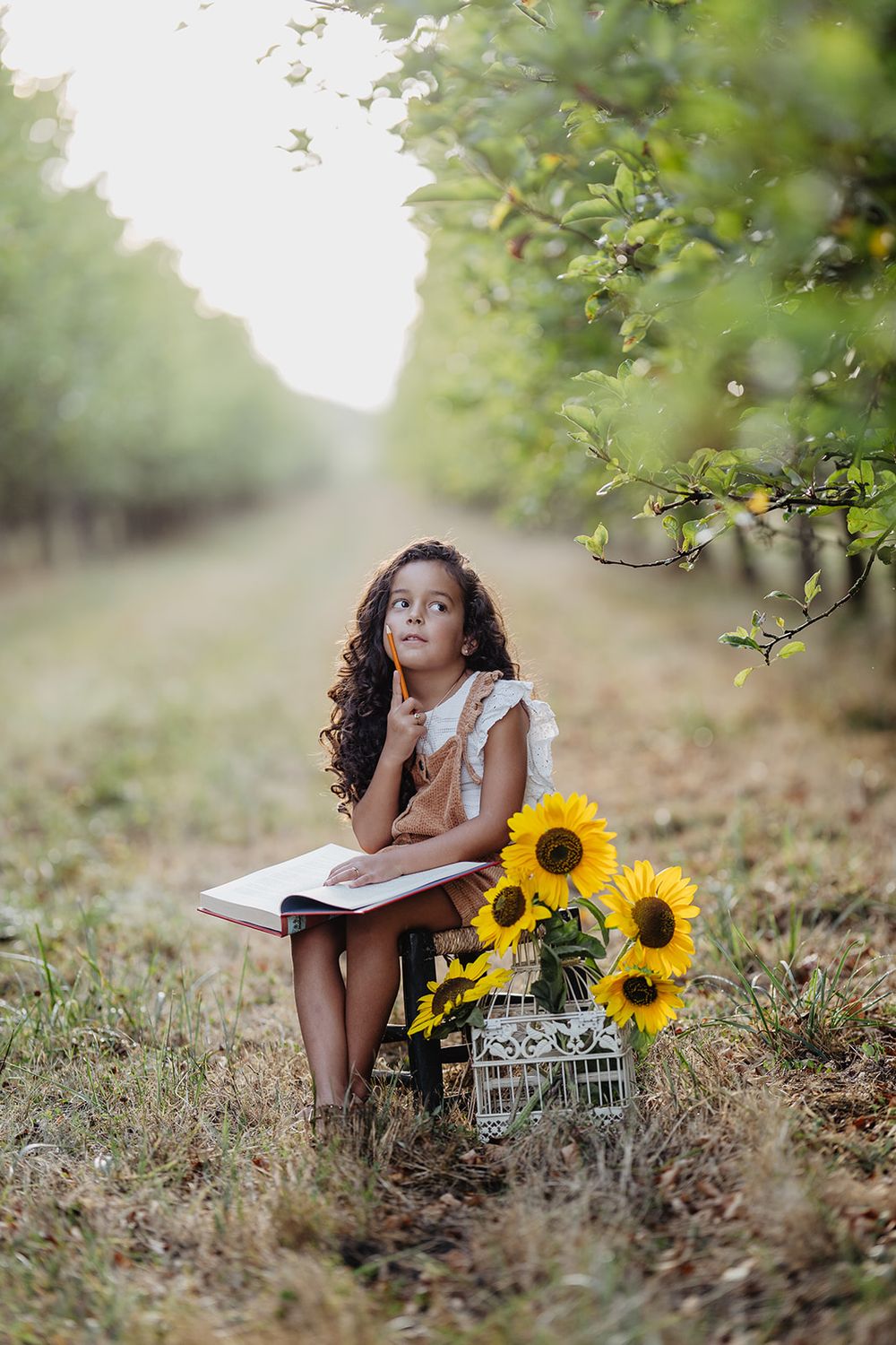 Young girl sitting outdoors in an orchard with a book and sunflowers, looking up thoughtfully.