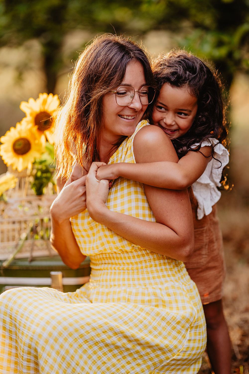 A woman in a yellow dress hugs a smiling young girl outdoors, with sunflowers in the background.