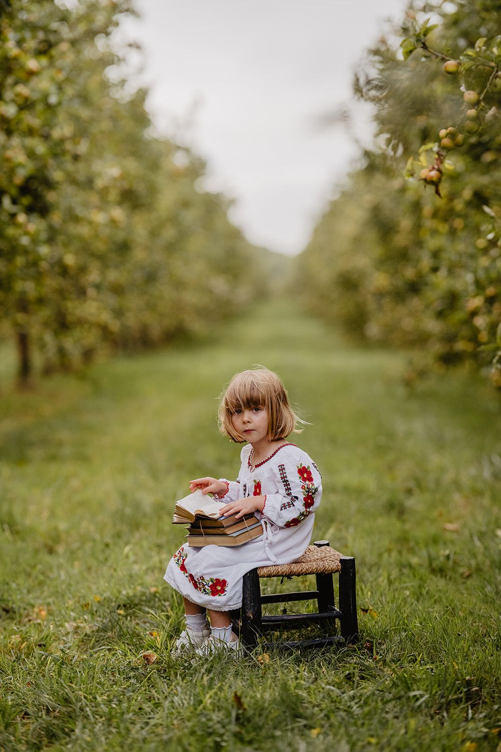 A young blonde girl in an embroidered dress reads a book while seated on a stool in an apple orchard.