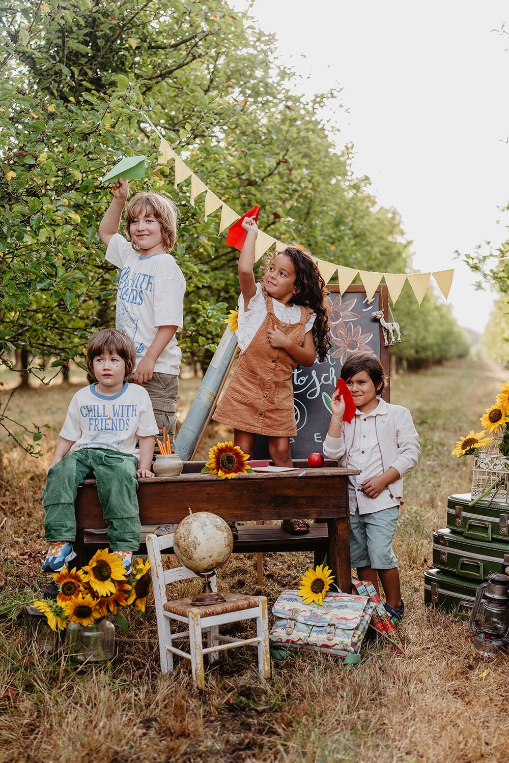 Four children playing outdoors around a vintage desk with sunflowers, a globe, and a chalkboard in a lush orchard setting.