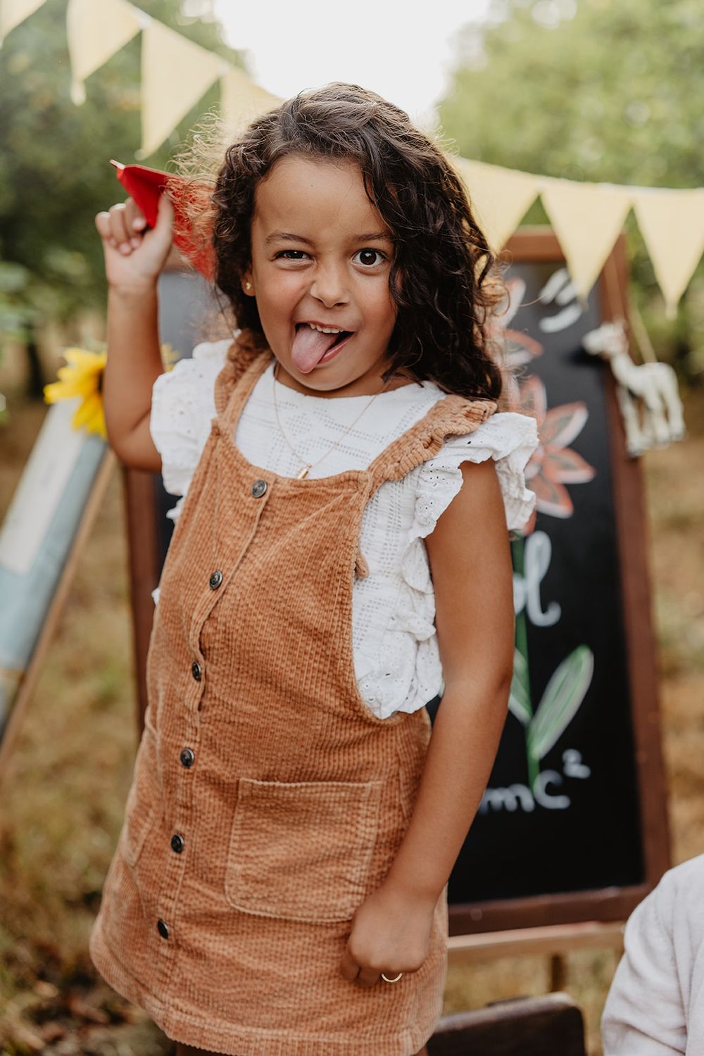 A young girl in a brown corduroy dress sticks out her tongue playfully outdoors near a chalkboard sign.