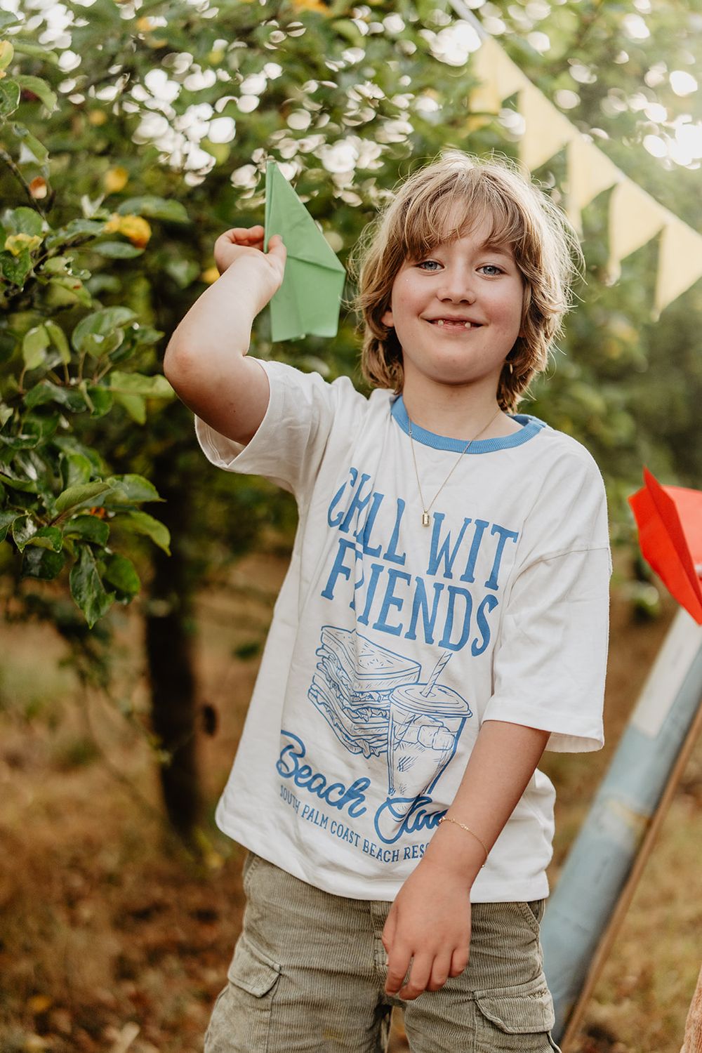 Smiling boy holding a green paper airplane outdoors, wearing a white Chill With Friends Beach Life graphic tee.