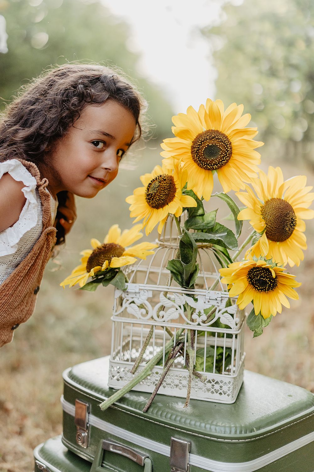 A young girl admires bright yellow sunflowers arranged in a vintage white birdcage atop a green suitcase outdoors.