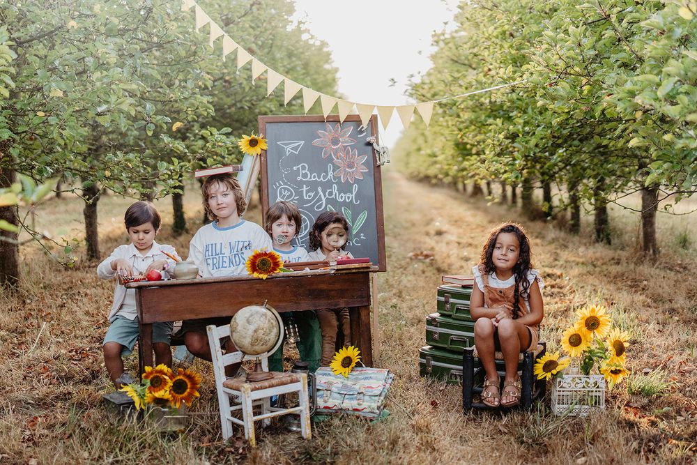 Four children pose around a chalkboard desk with sunflowers in an orchard for a back-to-school photo shoot.