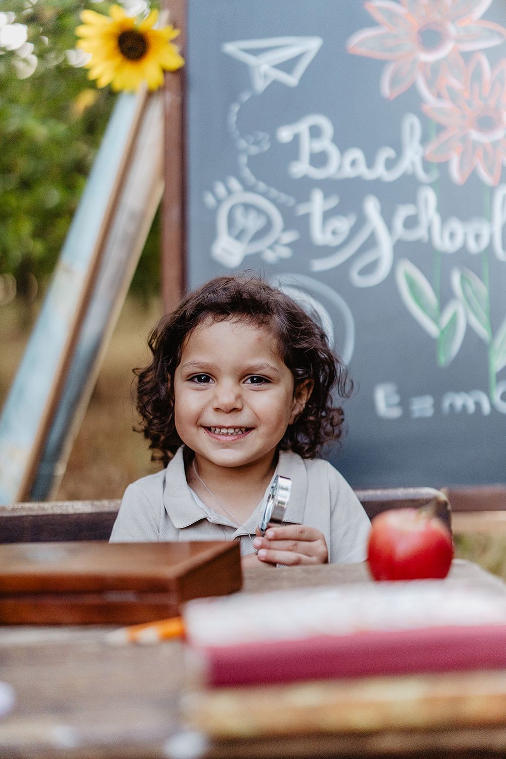 Smiling child with curly hair sitting at a desk outdoors in front of a Back to School chalkboard with an apple and books.