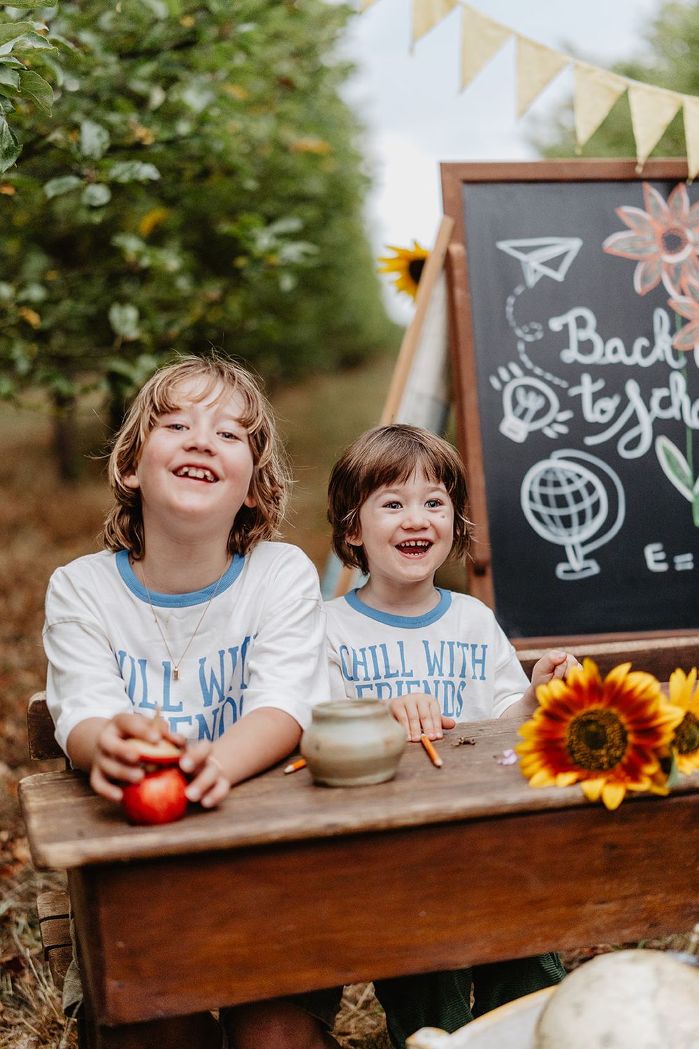 Two smiling children in matching shirts sit at a wooden desk with a back-to-school chalkboard and sunflowers outdoors.
