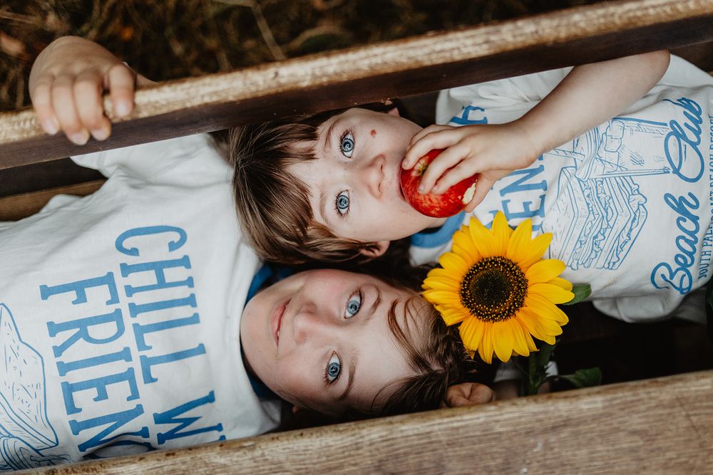 Two children with blue eyes lying in a wooden box, one holding a sunflower and apple, wearing matching graphic tees.