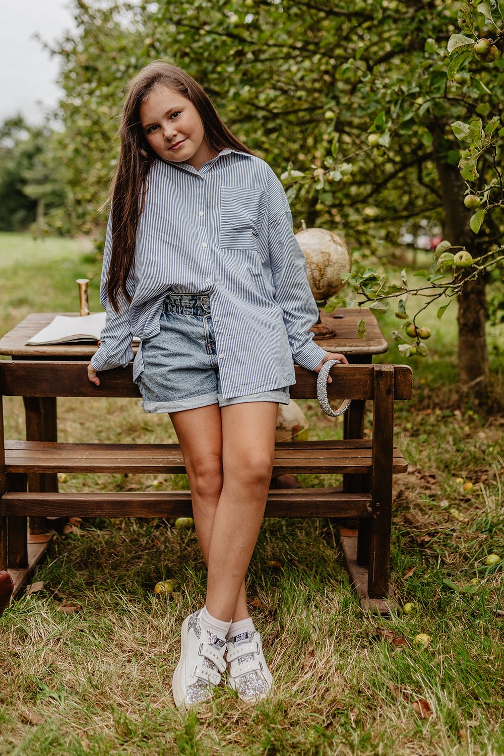 A teenage girl in a striped shirt and denim shorts leans against a wooden bench in a green orchard.