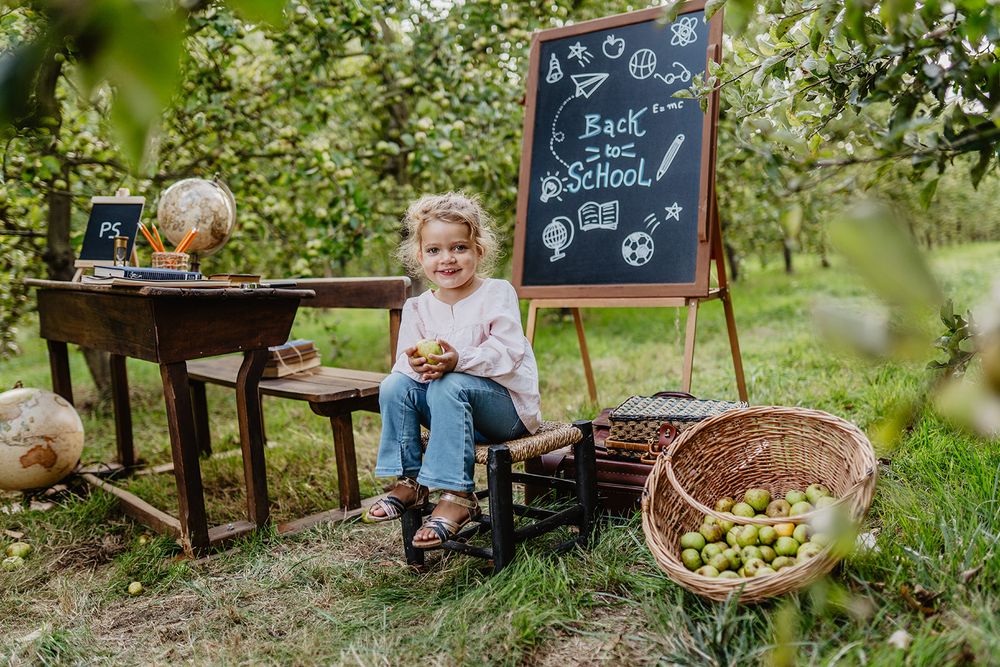 A smiling blonde girl sits near a Back to School chalkboard easel in a lush apple orchard setting.