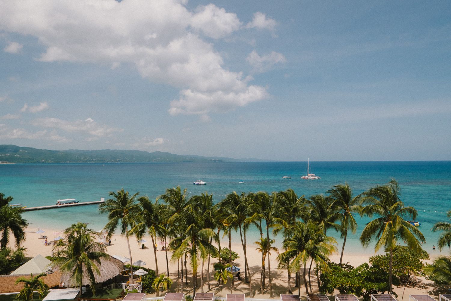 Panoramic view of a tropical beach resort with palm trees, white sand beaches, and turquoise ocean waters beneath blue skies.