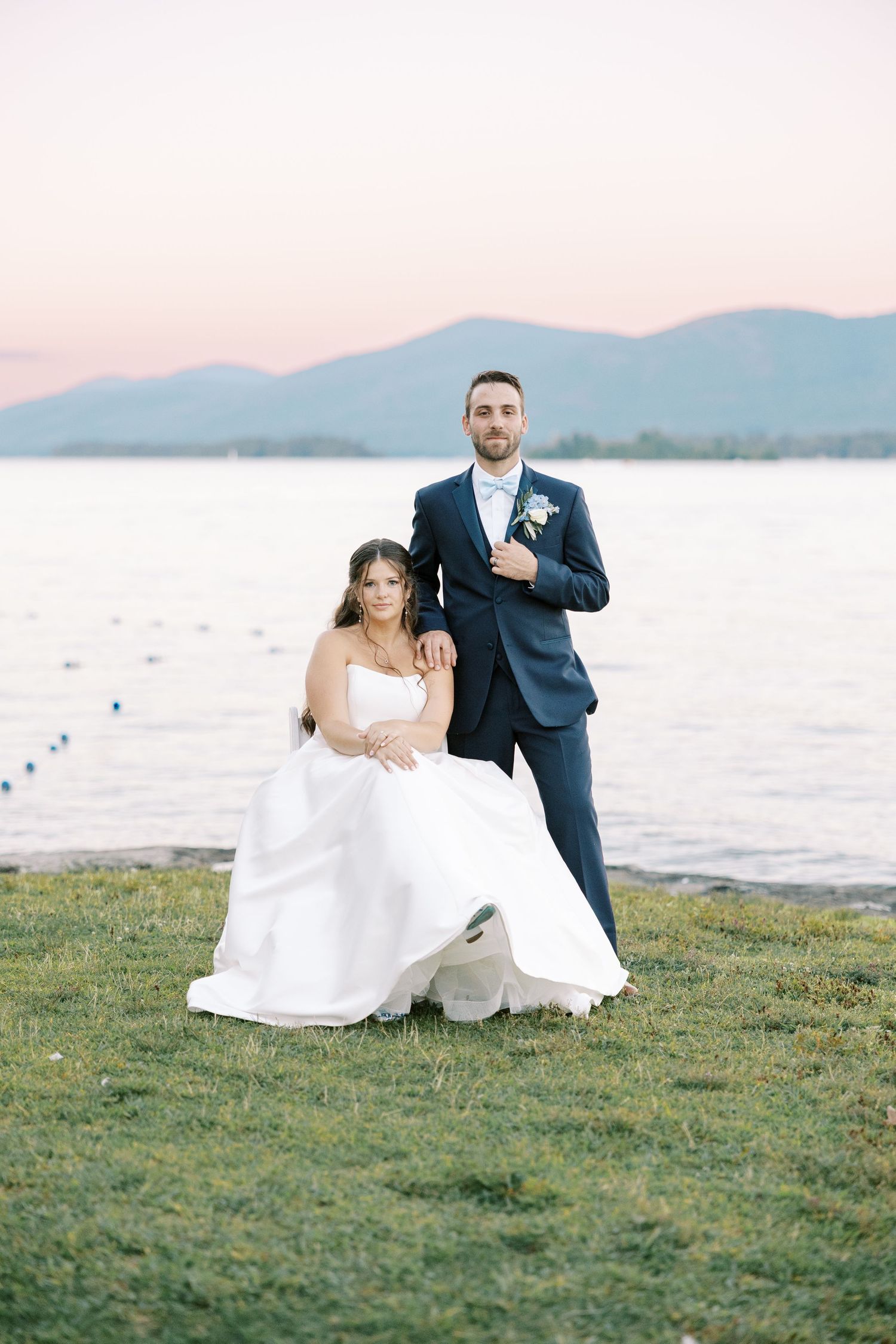 A couple in wedding attire sits on grassy lawn next to tranquil water at sunset during outdoor reception.