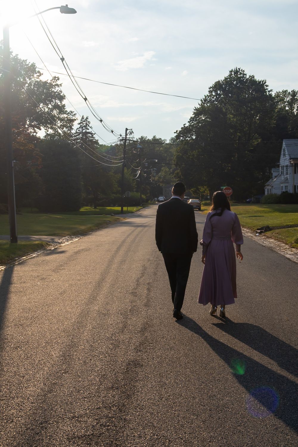 Couple walking down a sunny suburban street lined with trees, man in dark suit and woman in lavender dress.