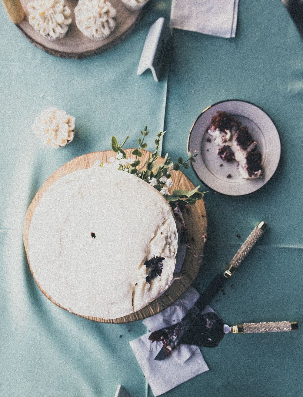 A white frosted cake on a wooden board with a slice removed, displayed on a teal tablecloth with flowers and a knife.
