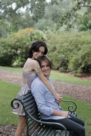 Two people share an intimate moment while sitting on a decorative park bench surrounded by greenery.