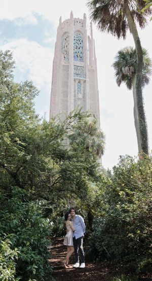 A couple stands beneath a towering Gothic bell tower surrounded by palm trees and greenery.