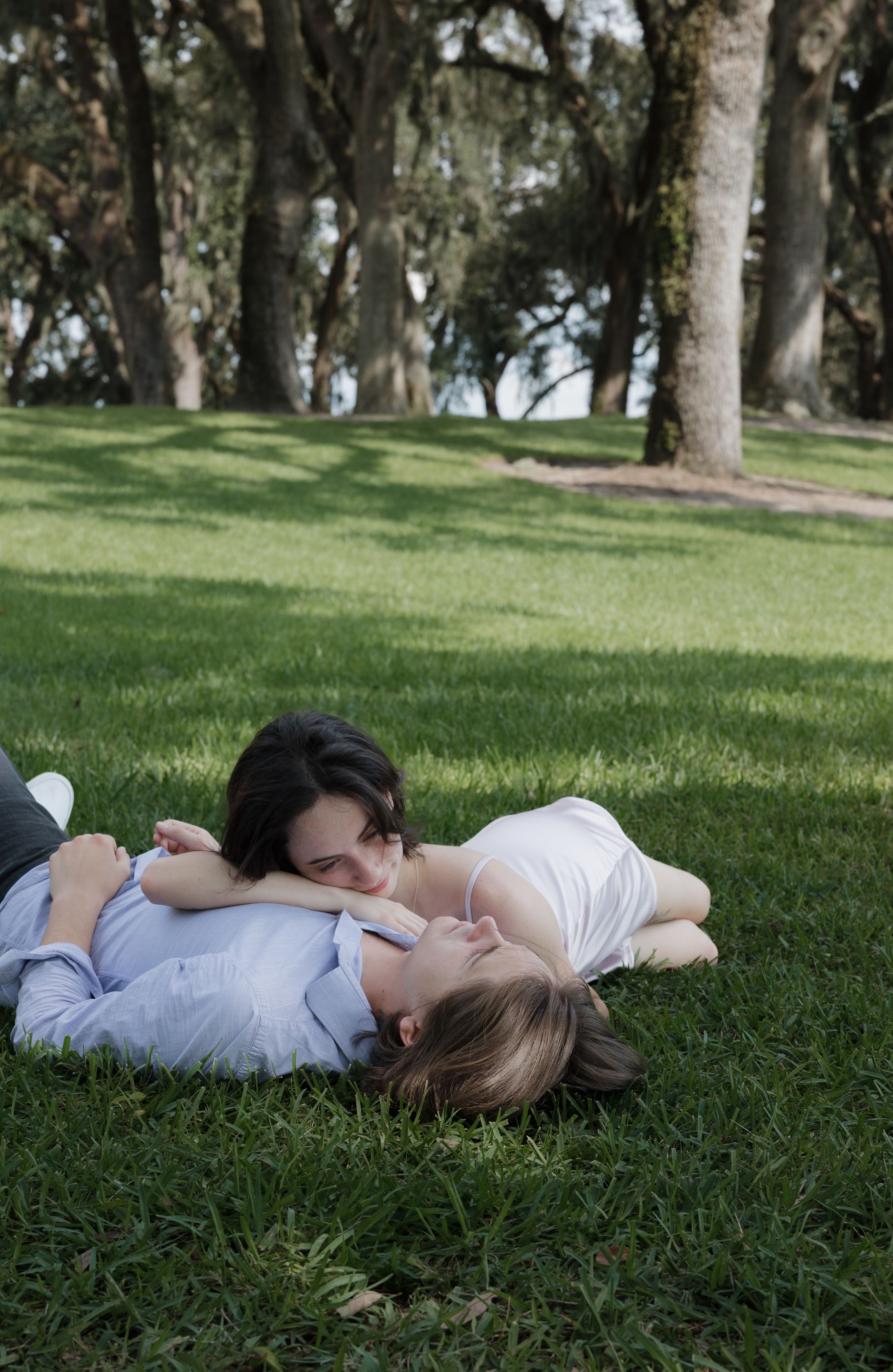 A couple lies together on grass beneath oak trees.
