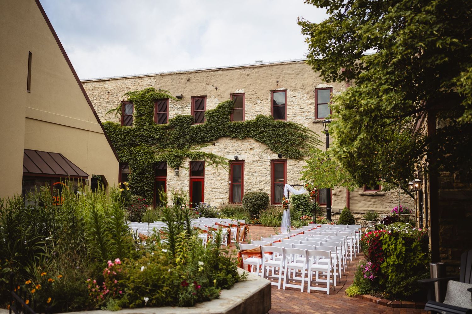 Outdoor wedding ceremony setup at a rustic stone venue with white chairs and flower decorations along a garden path.