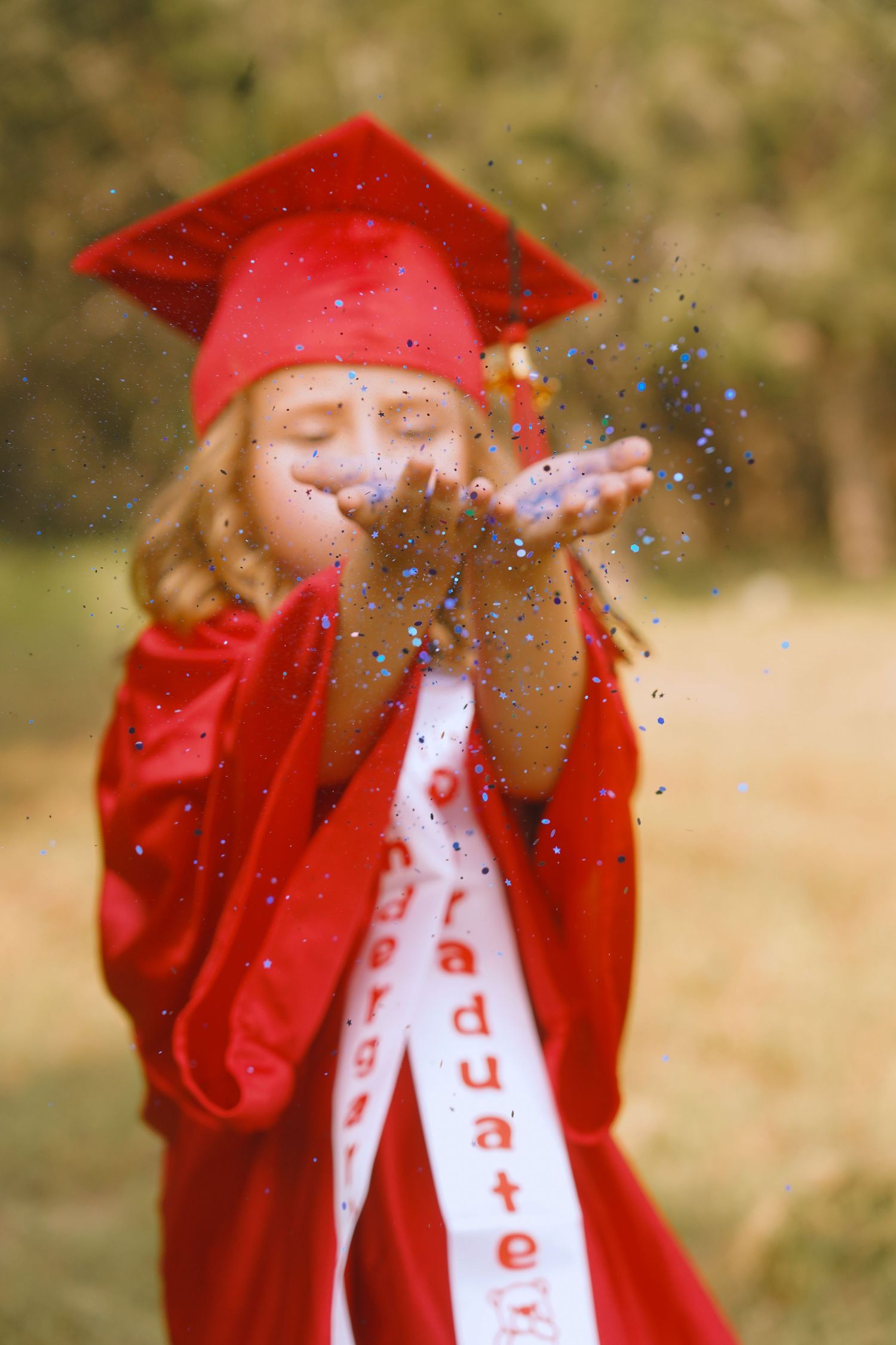 Graduate in red cap and gown blowing glitter in a celebratory outdoor portrait.