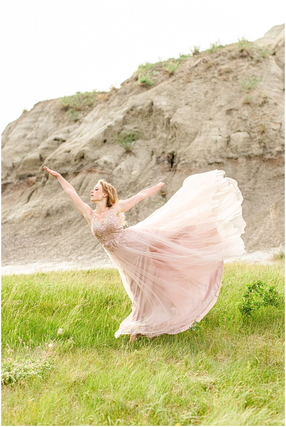 Ethereal dance pose in a flowing pink tulle gown against rocky cliffs and green grass.