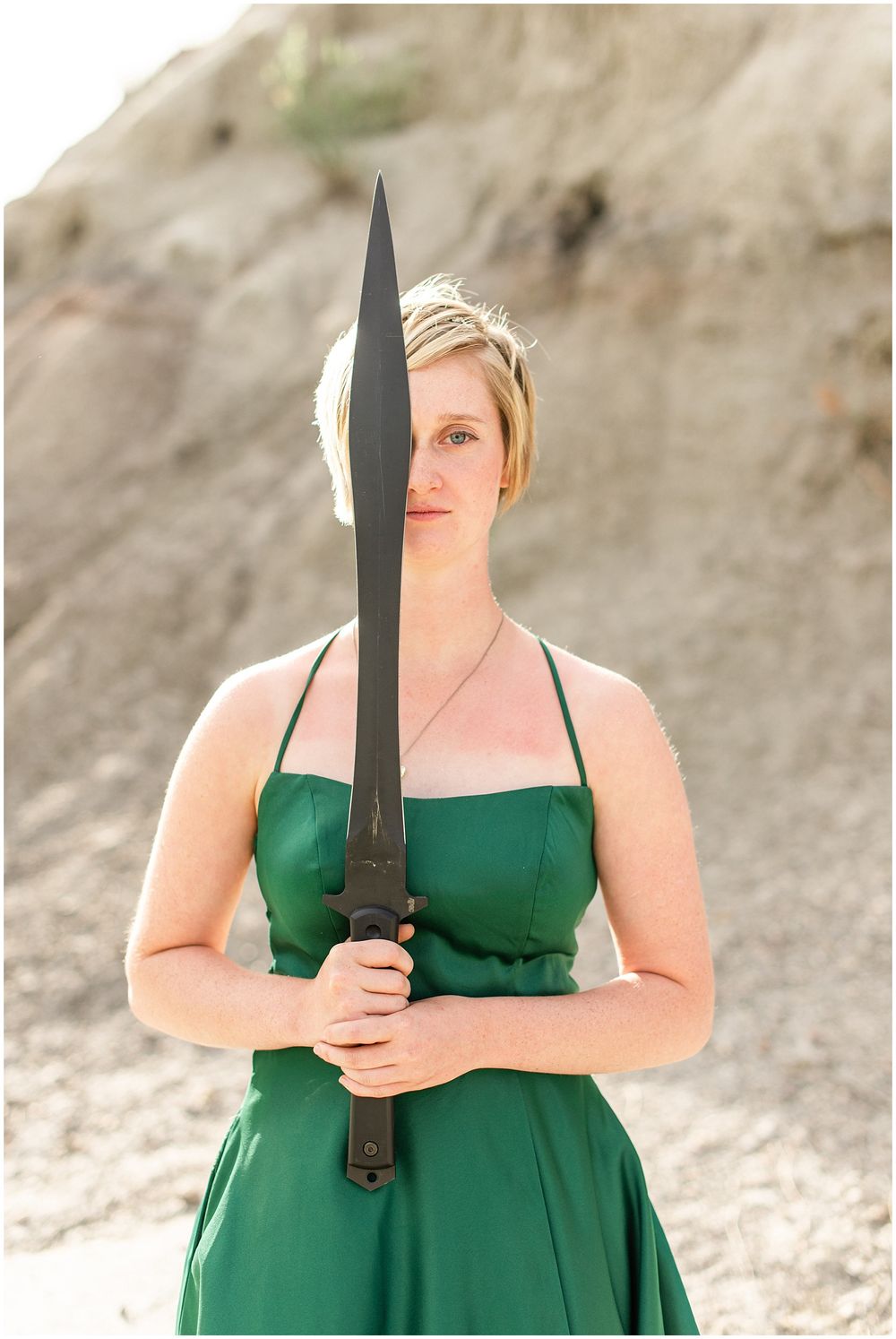 Figure in emerald green dress poses dramatically with sword against rocky backdrop.