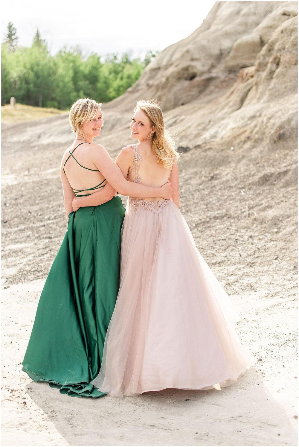 Two friends embracing in elegant formal dresses against rocky landscape.