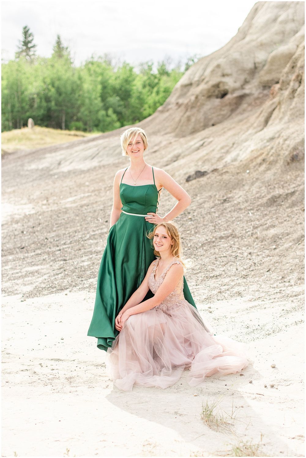 Two people in contrasting green and pink formal dresses pose against rocky terrain.