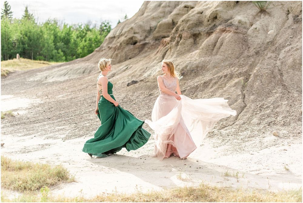 Two people twirling in flowing formal dresses against rocky landscape.