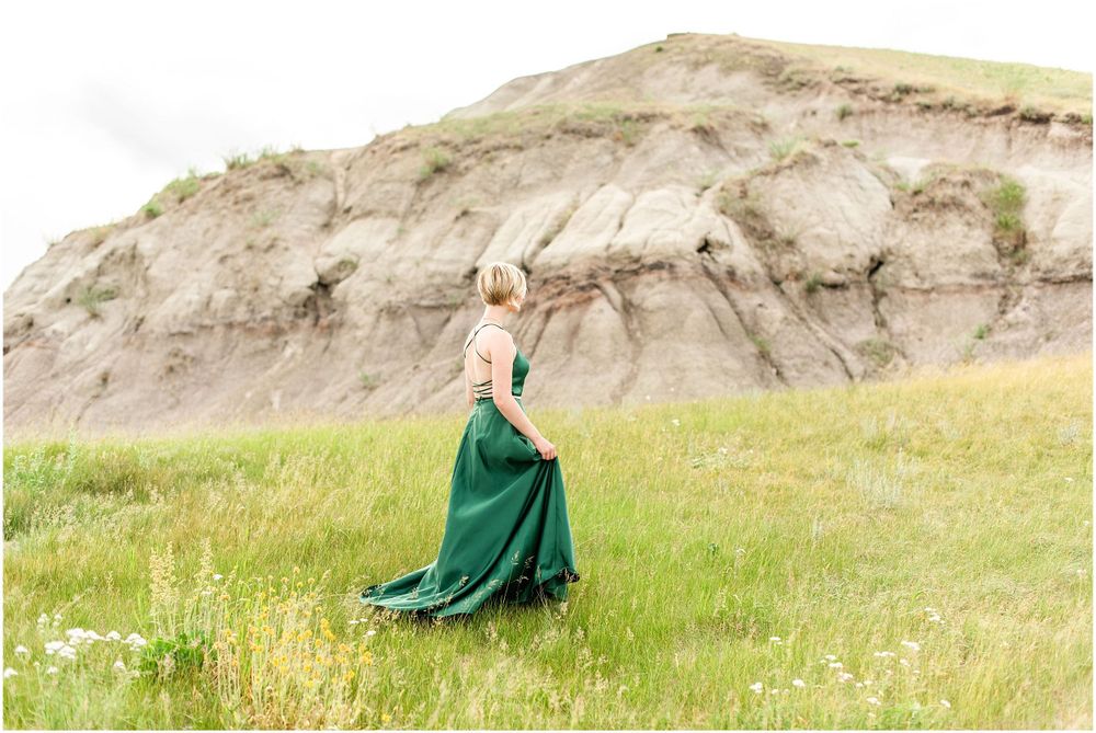 Person in emerald green gown poses dramatically on grassy hillside.