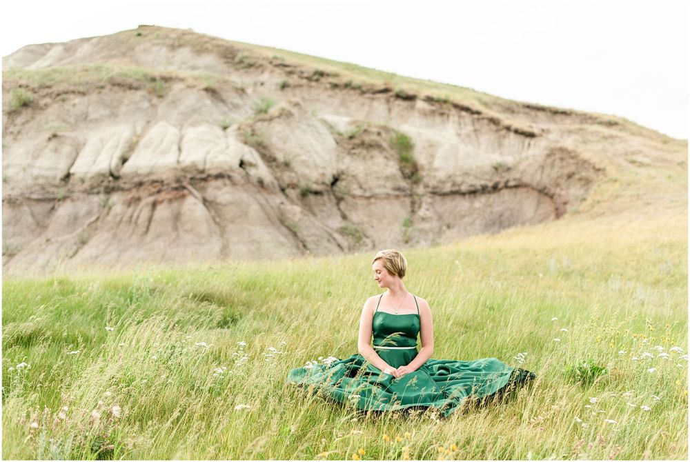 Elegant emerald green gown flows in a grassy field against rugged cliffs and hills.