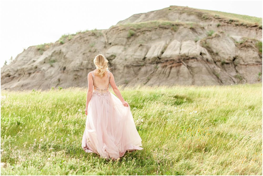 Soft pink gown flows gracefully in meadow with dramatic cliff backdrop.
