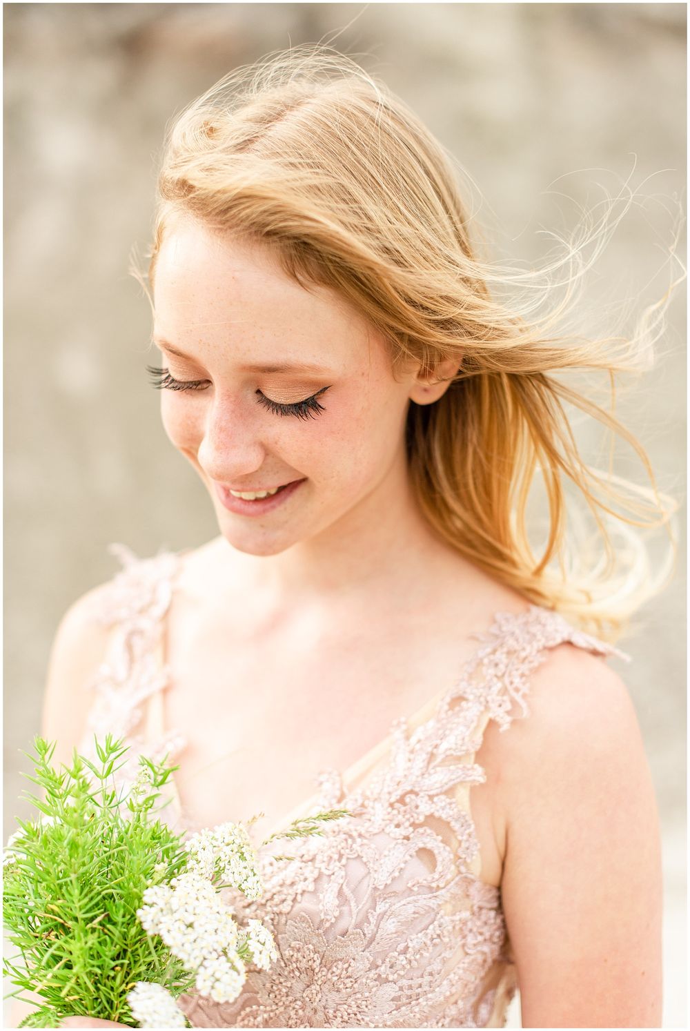 A portrait in blush pink lace gown with windswept hair and natural bouquet against soft background.