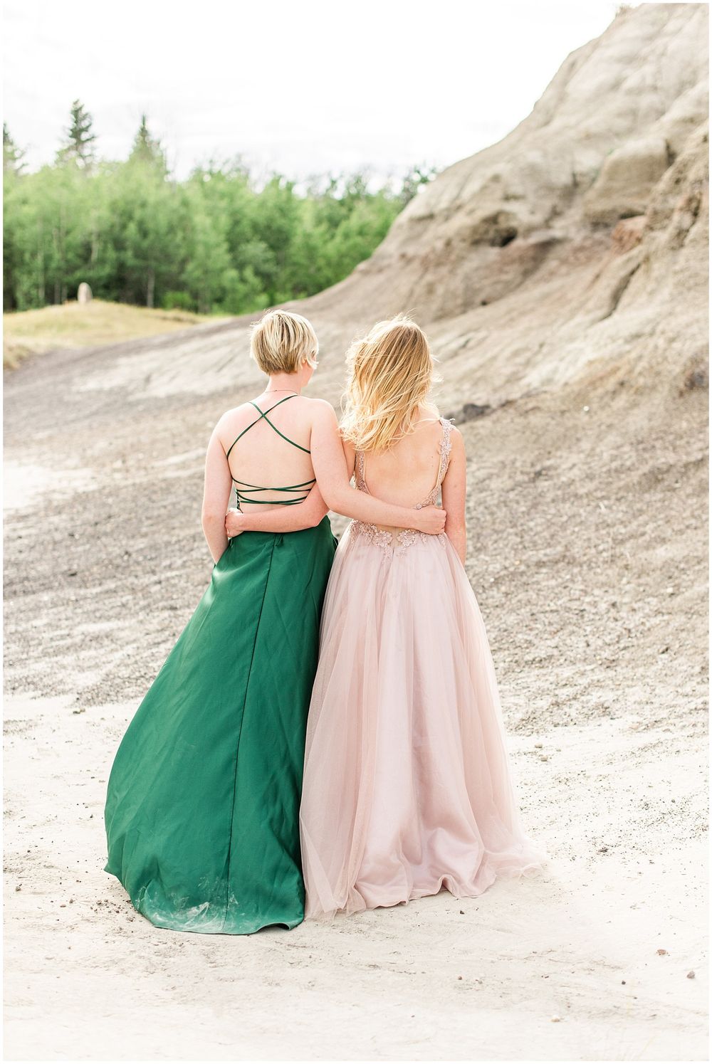 Two figures in contrasting pink and green formal dresses embrace against rocky landscape.
