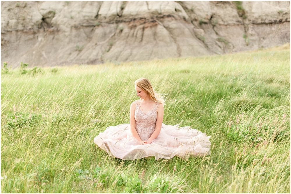 A woman in a pink dress sits gracefully in tall grass with rocky cliffs in the background.