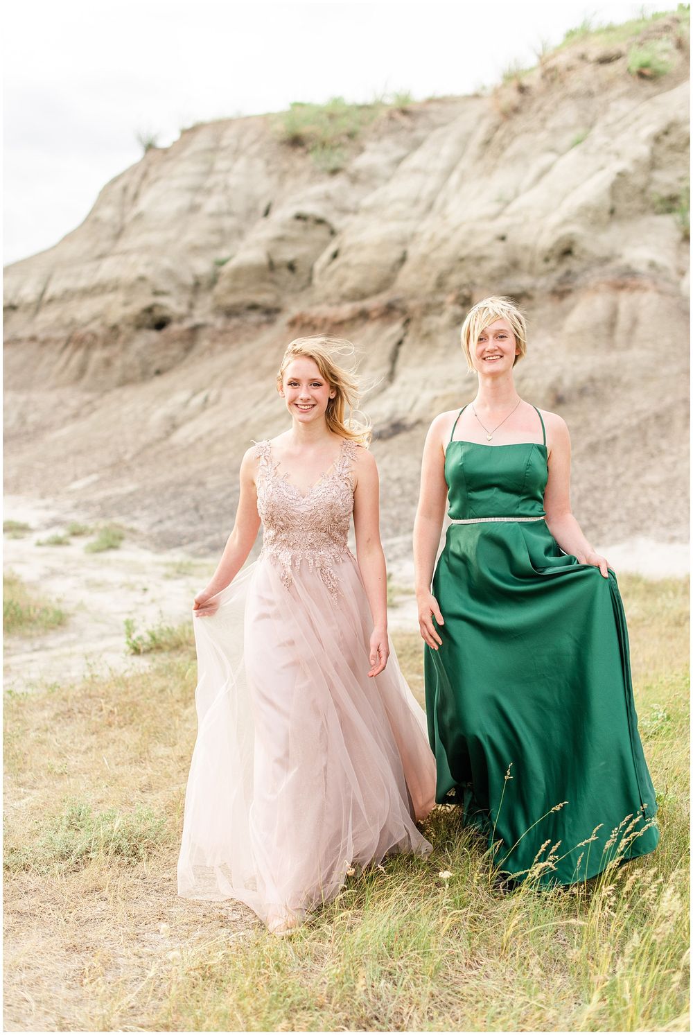 Two figures in pink and emerald formal gowns pose together against rocky cliff background.