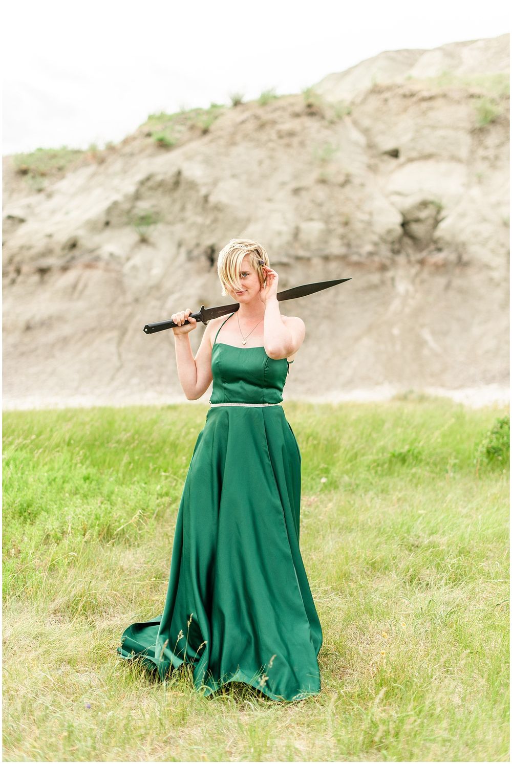 A figure in a flowing emerald gown holds an umbrella against a backdrop of rugged cliffs.
