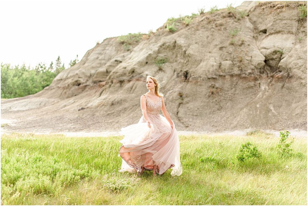 A person in a blush gown twirls in a grassy meadow with rocky cliffs behind.