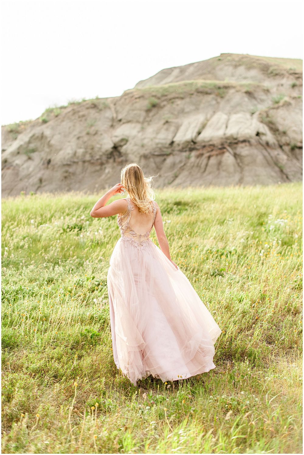 Person in flowing pink gown stands in grassy field with dramatic cliffs in background.
