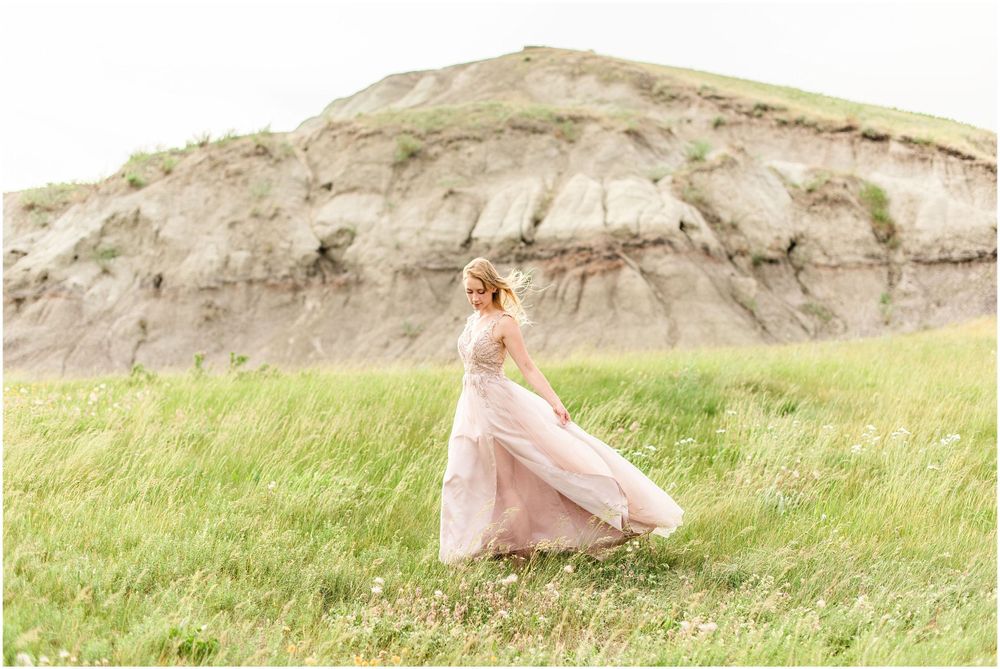 A person in a flowing pink gown stands against windswept grass and rocky terrain.
