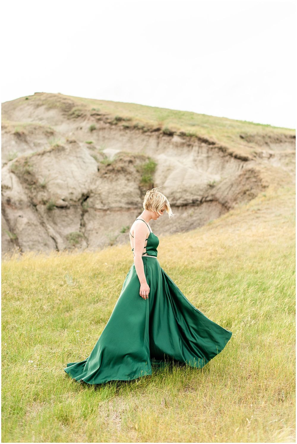 A figure in an emerald ball gown walks gracefully across a grassy hillside.