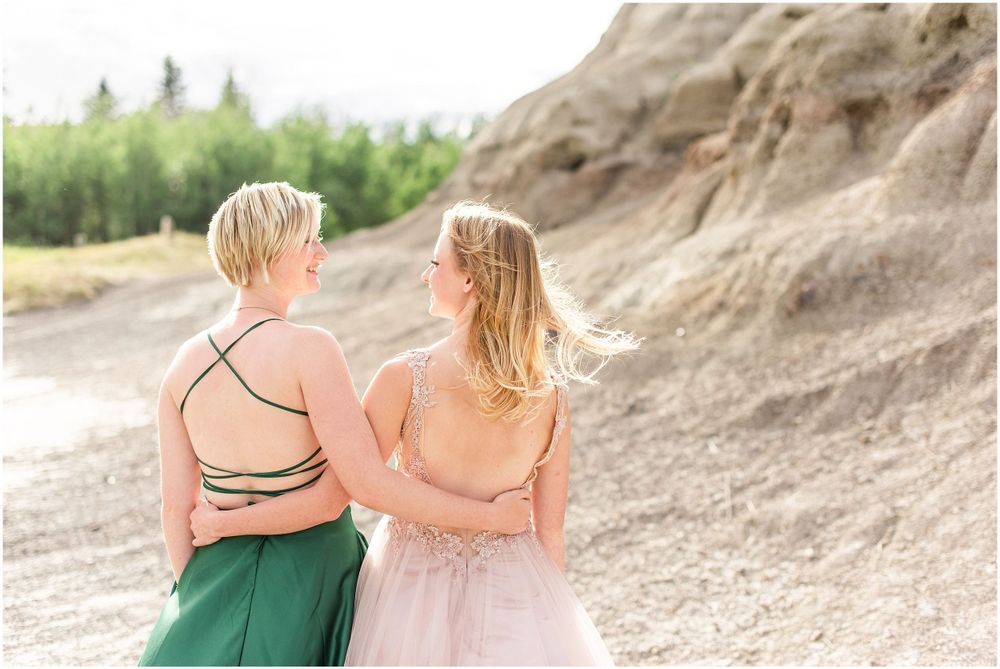 Two people in elegant dresses pose together against rocky cliffs in a romantic photoshoot setting.