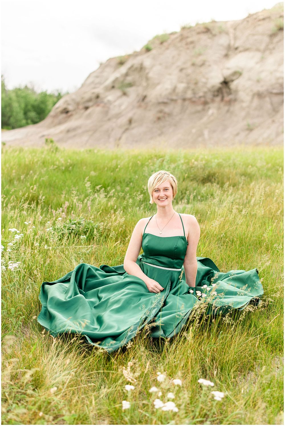 A flowing emerald dress spreads across wild grass with rocky cliffs in the background.