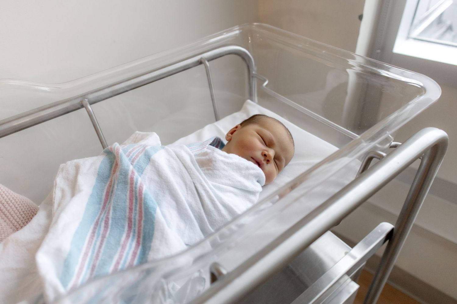 A sleeping newborn baby swaddled in a striped blanket rests peacefully in a hospital bassinet.