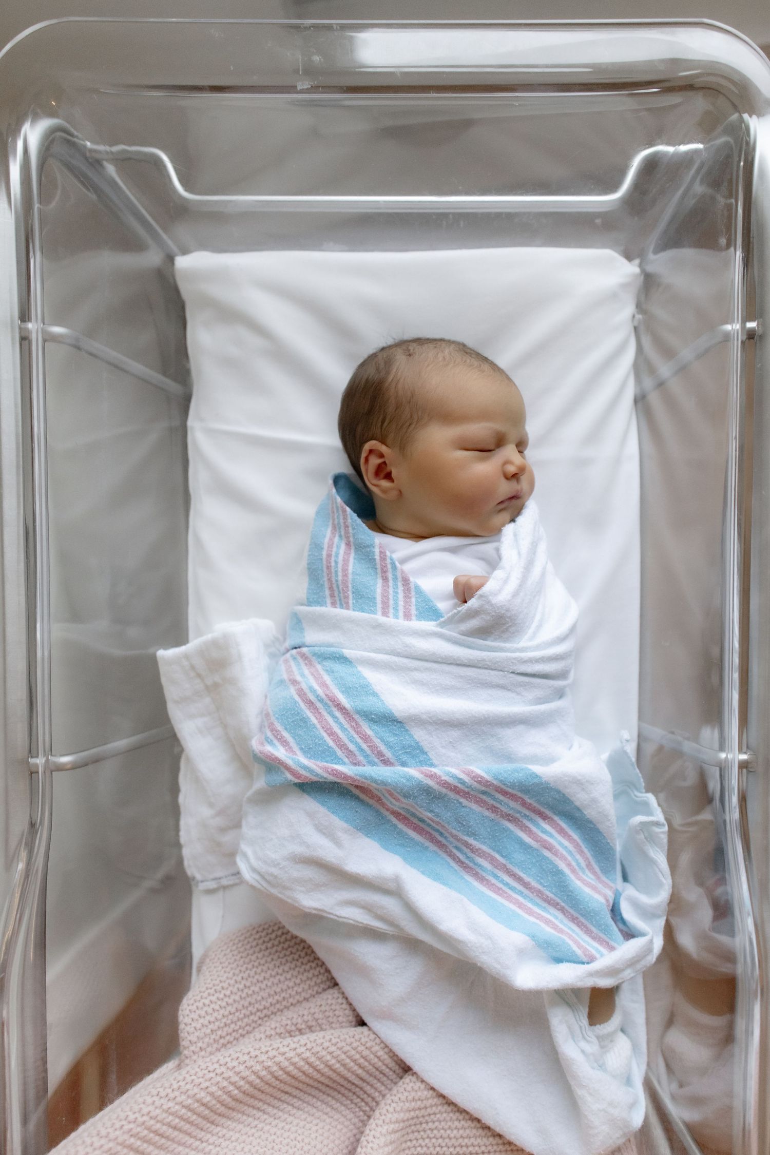 Sleeping newborn baby swaddled in white and blue striped blanket lying in clear hospital bassinet.