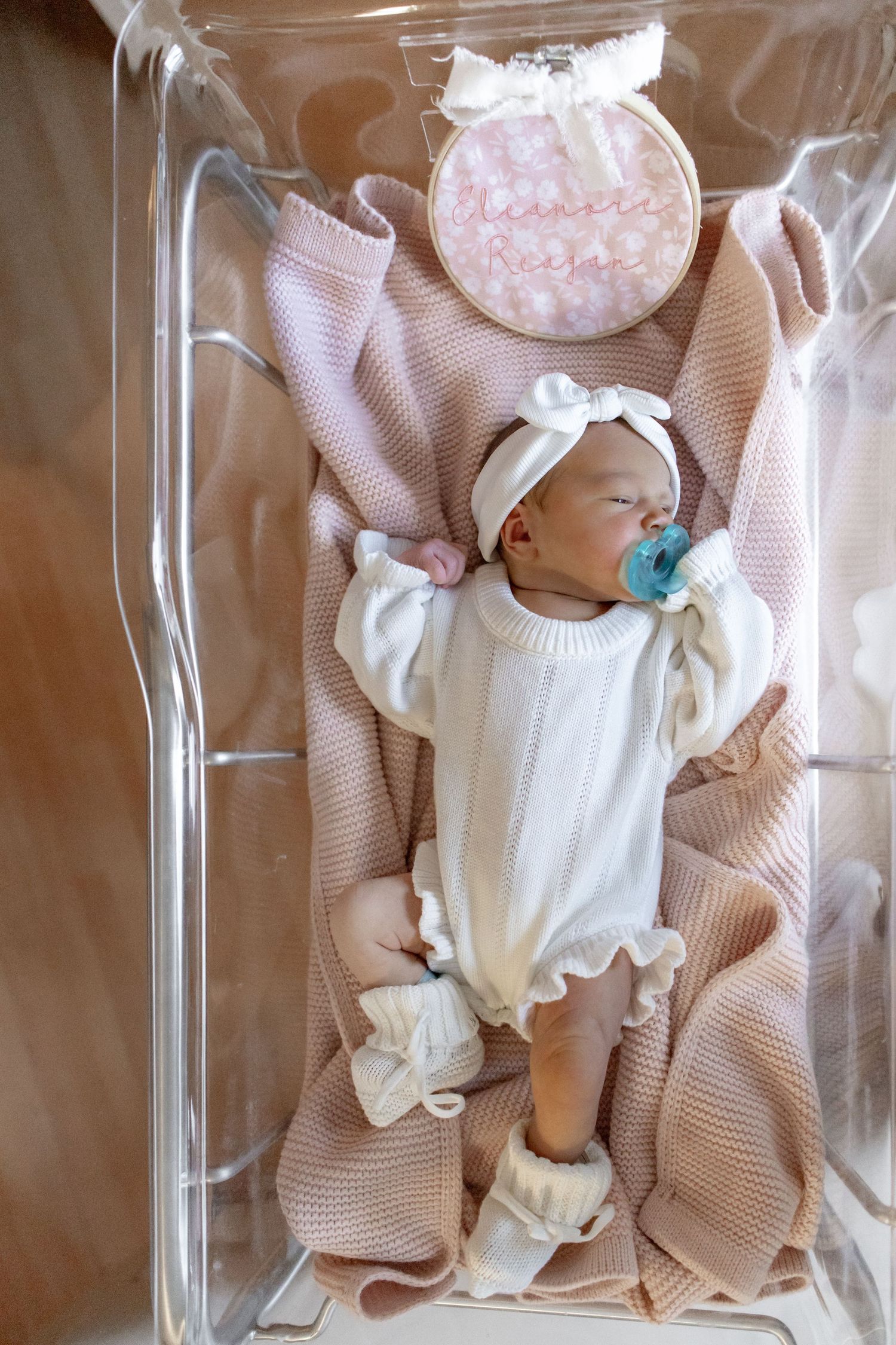 Close-up sequence of a newborn baby in white onesie resting on a tan textured blanket under clear plastic nursery bassinet.