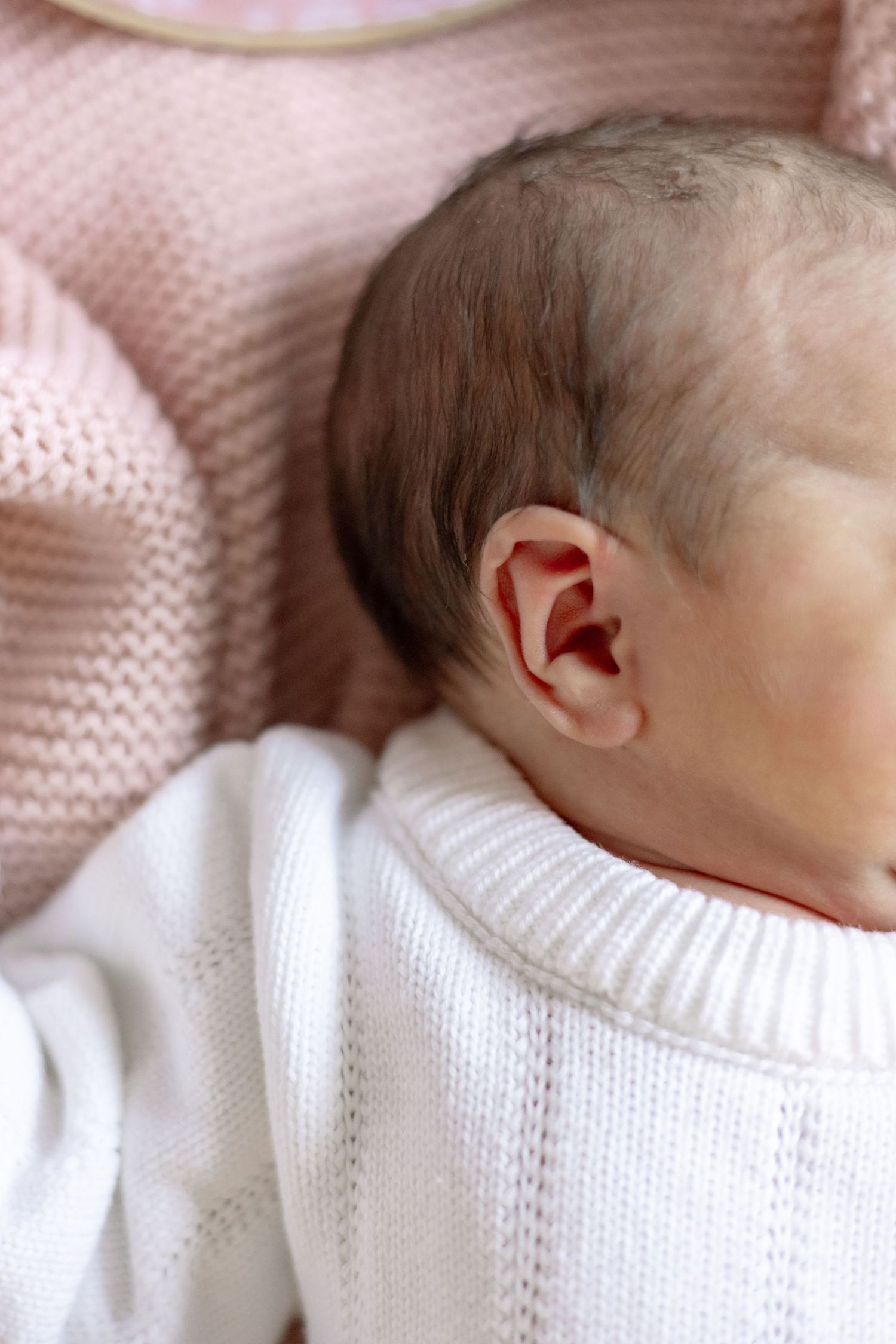 Sleeping newborn baby wearing white knit sweater rests peacefully on pink blanket.